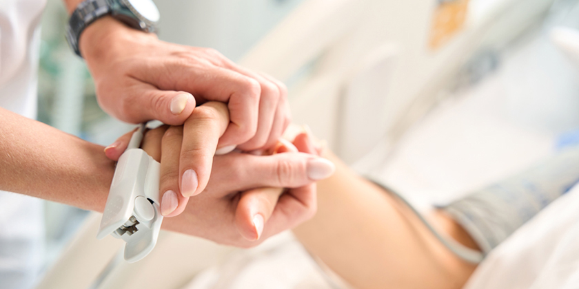 A close-up of a doctor's hands holding a patient's hand at a hospital bed.