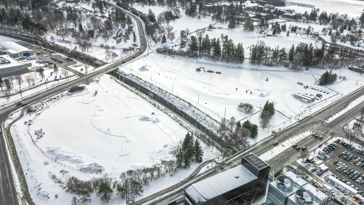 An aerial view in winter of the snow-covered construction site where the parking garage for The Ottawa Hospital’s new campus will be built. There are a few pieces of construction equipment, some trailers and a tractor on the site.