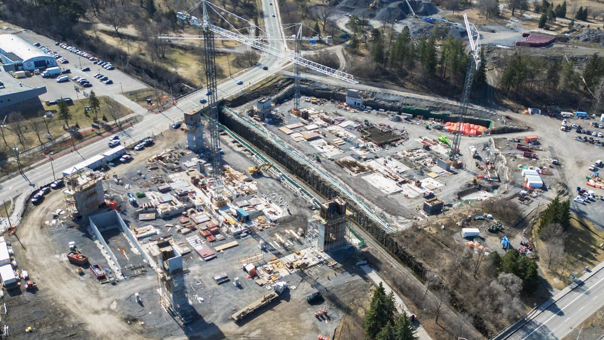 An aerial view in early spring of the construction site for the parking garage at The Ottawa Hospital’s new campus. Three large tower cranes are on the site. Large concrete structures like stairwells, elevator shafts and an entrance ramp for vehicles are partially built, extending vertically up from the ground level.