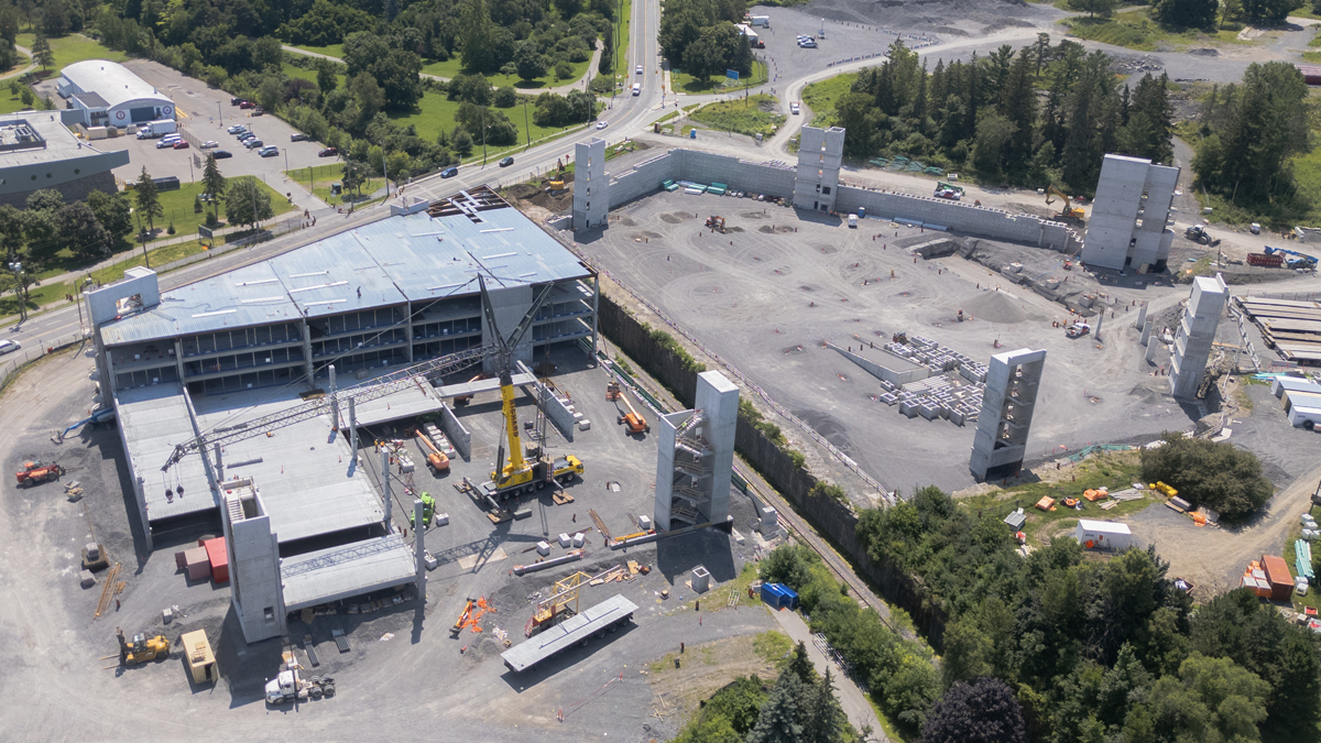 An aerial view in summer of the partially constructed parking garage at The Ottawa Hospital’s new campus. A large yellow crane is on the site, and multiple levels on one half of the parking garage have been built. An unfinished roof covers the portion of the garage that is complete.