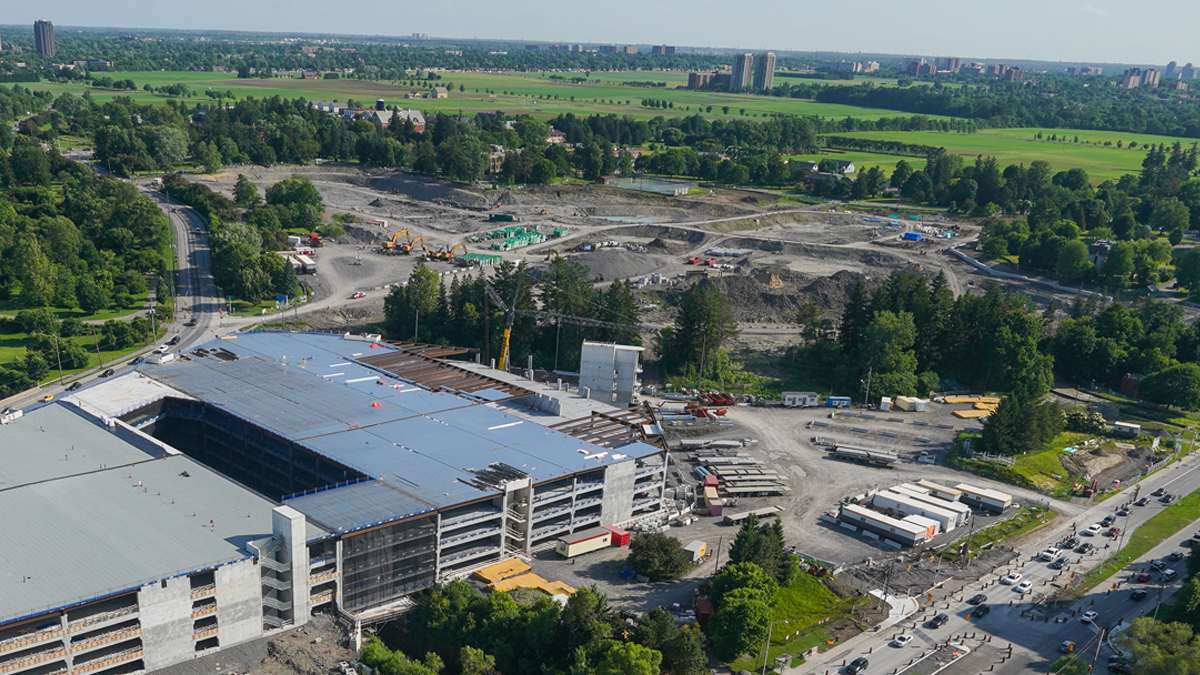 An aerial view of the construction site of the new hospital. A multi-level parking garage is under construction at lower left, and a large area of land is being excavated at top centre. The site is surrounded by trees and grassy fields.