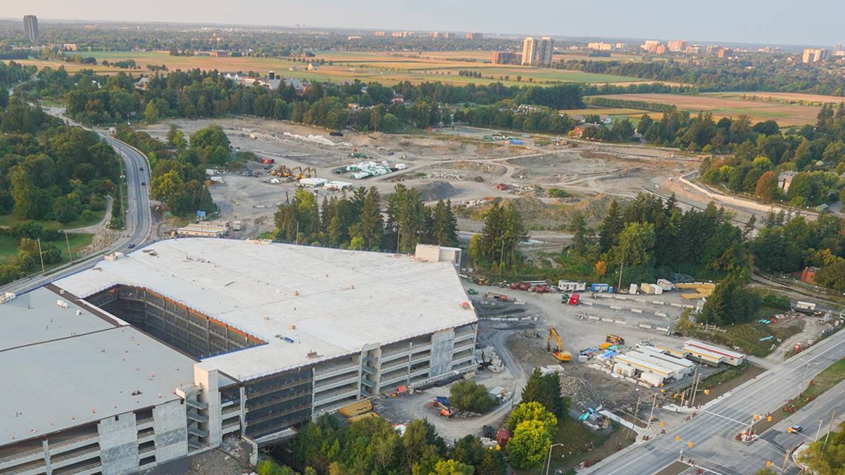 An aerial view of the construction site of the new hospital. A multi-level parking garage is under construction at lower left, and a large area of land is being excavated at top centre. The site is surrounded by trees and grassy fields.
