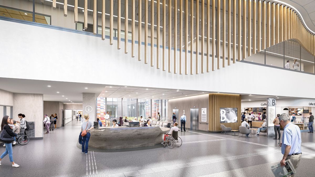 An artist rendering of the interior of the main entrance of a new hospital. At ground level in the lower half, there is a wide grey desk with hospital staff behind it. Two hallways lead in different directions on either side of the desk, and there are elevators and a coffee shop at the right. The top half shows an upper floor of the hospital. The busy scene shows people walking, seated and in wheelchairs at various locations.