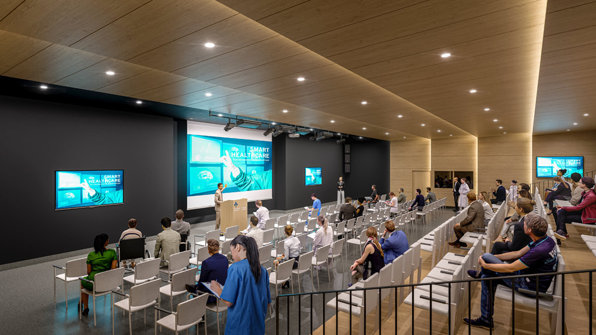 An artist rendering of the interior of an auditorium at a new hospital. A man is speaking at a podium; three large screens are on the wall behind him. There are multiple rows of chairs and various people seated throughout the large room.