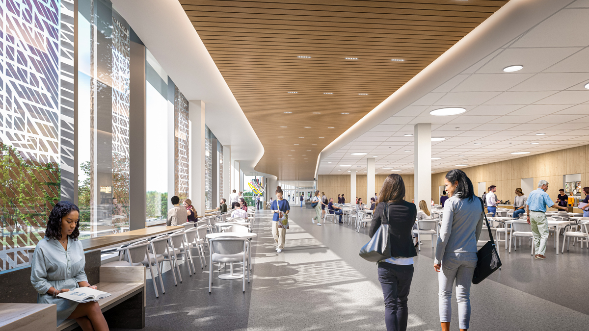 An artist rendering of the interior of a cafeteria at a new hospital. A long, open hallway from the middle foreground to the background. Floor-to-ceiling windows on the left of the hall fill the space with natural light. People are walking through the hall and sitting at tables and chairs on either side of the hall.